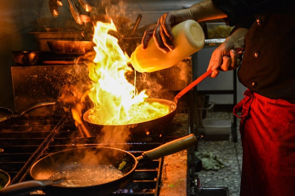 A cook tilting a pan over an open flame in a dark professional kitchen.