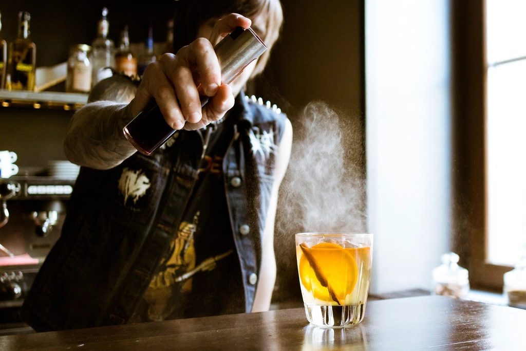 A bartender pouring a drink from a shaker at a daylit bar counter.