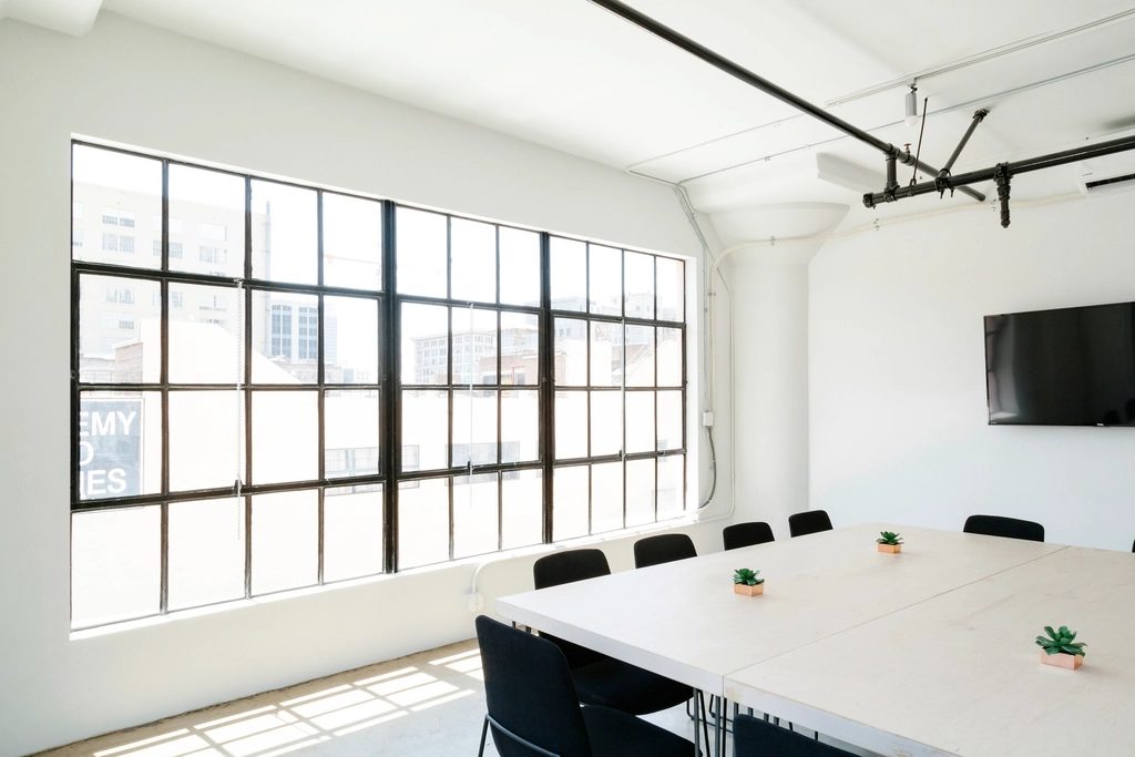 A bright white meeting room with a long table, black chairs and a wall of industrial windows.
